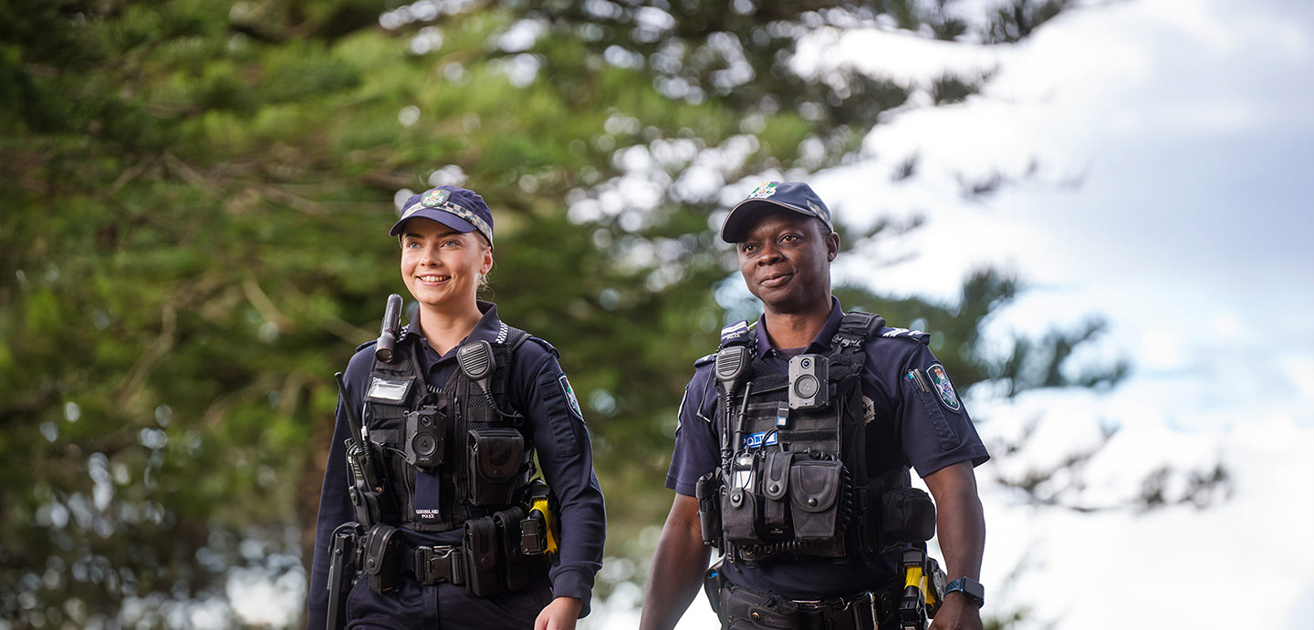 Two Queensland Police Service officers walking outside with a tree and patch of sky in the background.